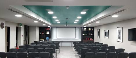 A conference room with rows of chairs, a projector screen, a screen displaying 'LOVE', and a shelf with books and decorative items.