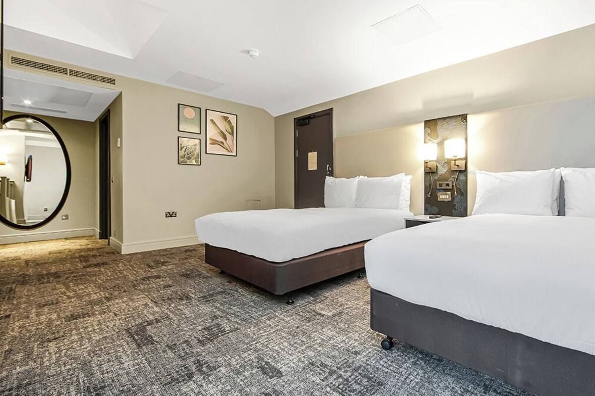 A hotel room with two beds, a round mirror, framed artwork, a door, and a carpeted floor.