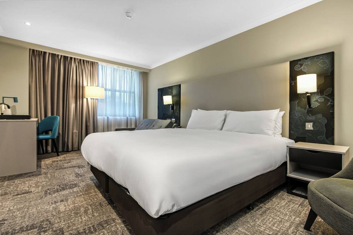 A hotel room with a large bed, a desk, a chair, and a nightstand with a lamp.