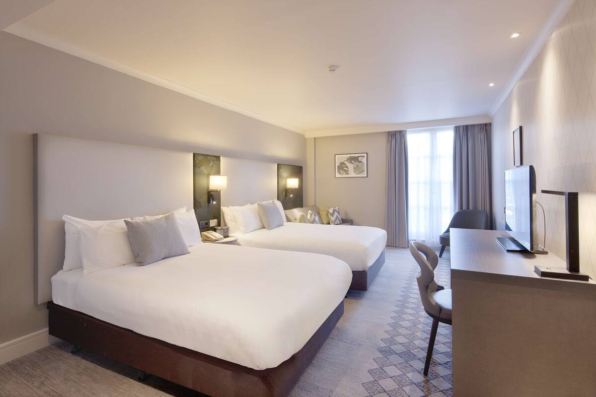 A hotel room with two beds, a desk, a chair, and a large window with curtains.