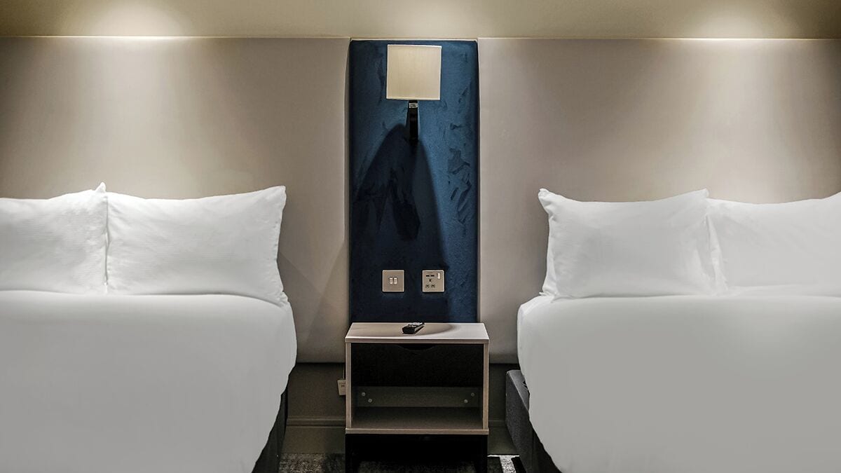 Een hotelkamer met twee eenpersoonsbedden, een nachtkastje met een lamp en een wandcontactdoos.