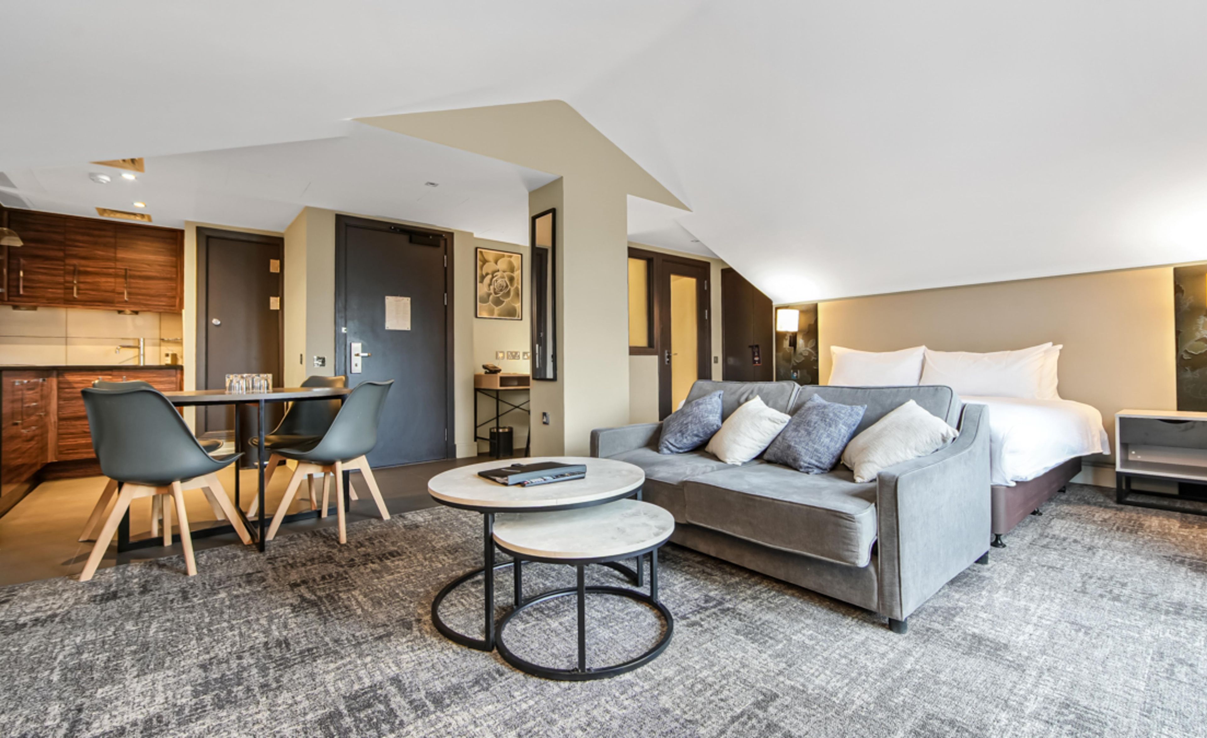 A modern hotel room with a sofa, a small round table, a dining area with chairs, a kitchen, and a bed with pillows.