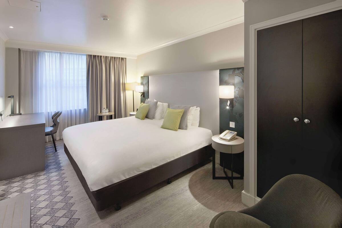 A hotel room with a large bed, bedside tables, a desk, a chair, and a wardrobe.