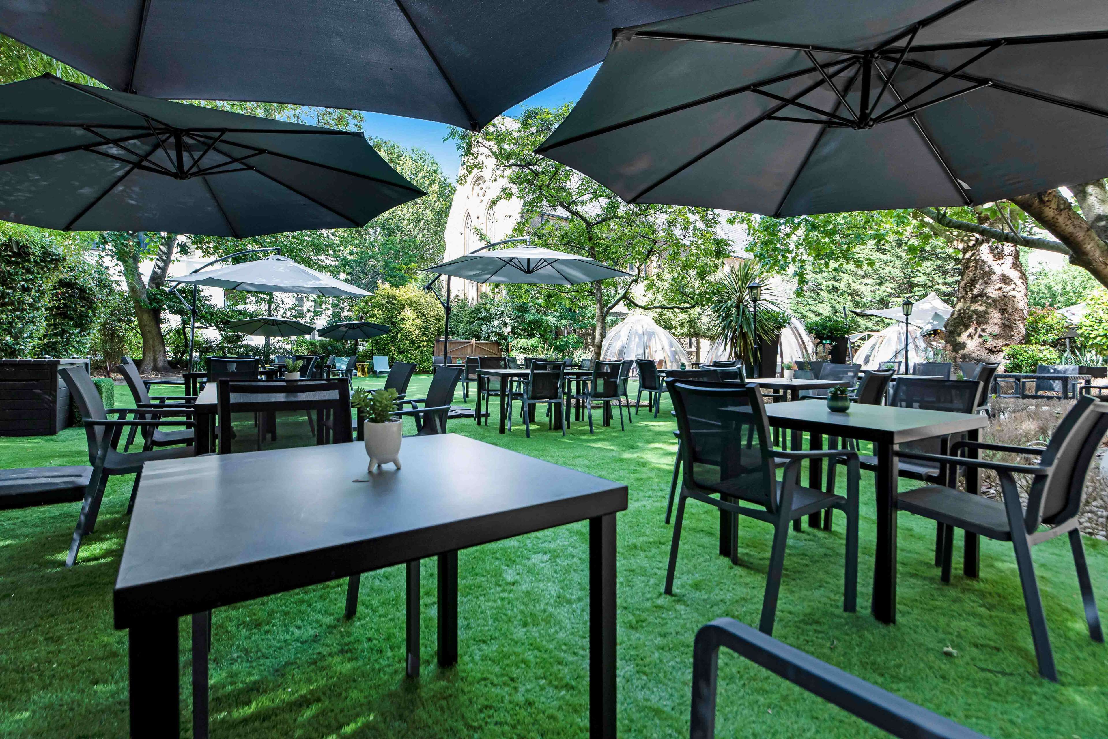 An outdoor patio area with tables and chairs under large umbrellas, surrounded by greenery and a building in the background.