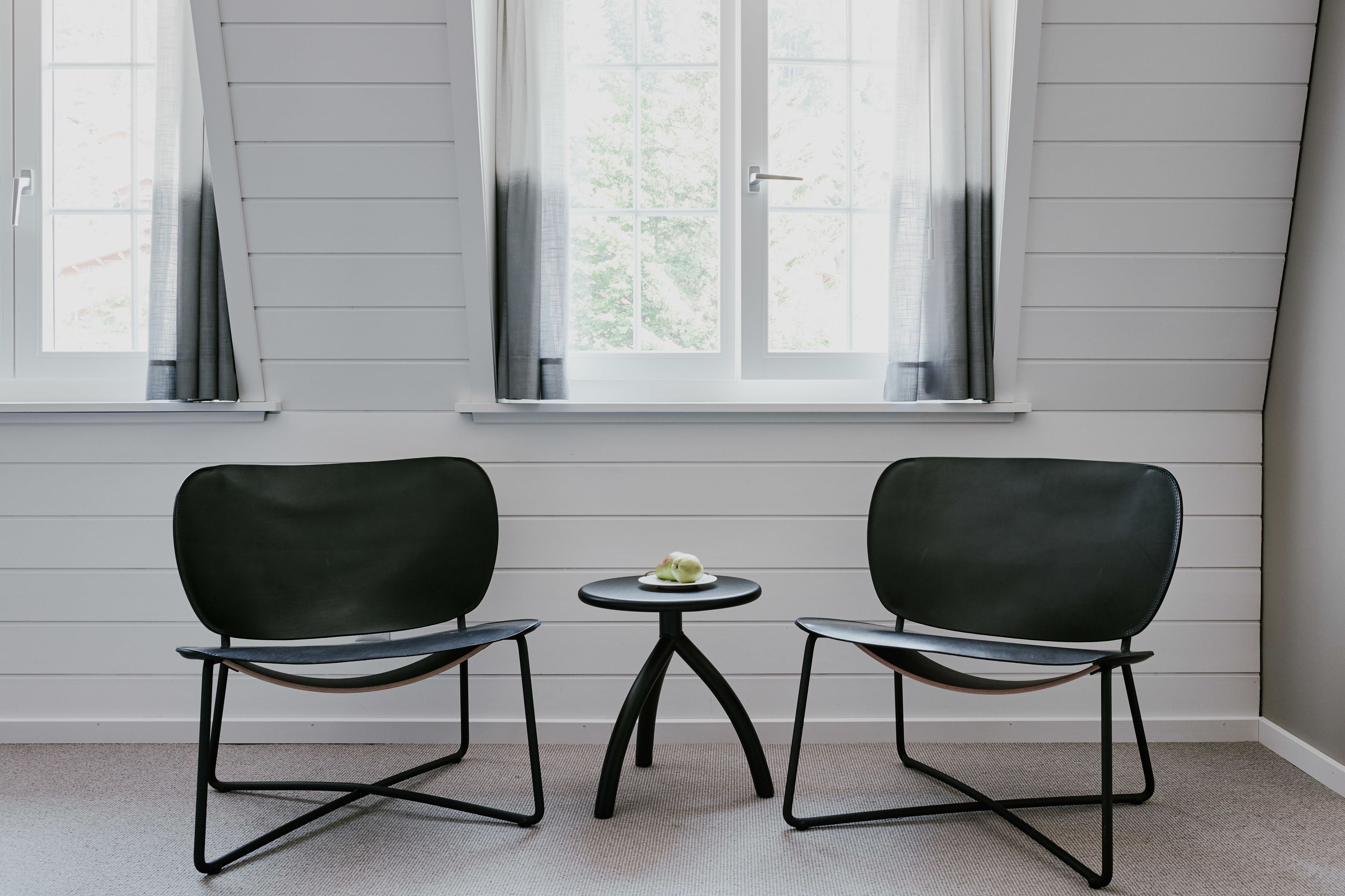 Ein Zimmer mit zwei schwarzen Stühlen und einem kleinen runden Tisch, auf dem ein weißes Objekt steht, gelegen zwischen zwei Fenstern mit weißen Rahmen und grauen Vorhängen.