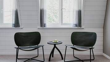 Ein Zimmer mit zwei schwarzen Stühlen und einem kleinen runden Tisch, auf dem ein weißes Objekt steht, gelegen zwischen zwei Fenstern mit weißen Rahmen und grauen Vorhängen.