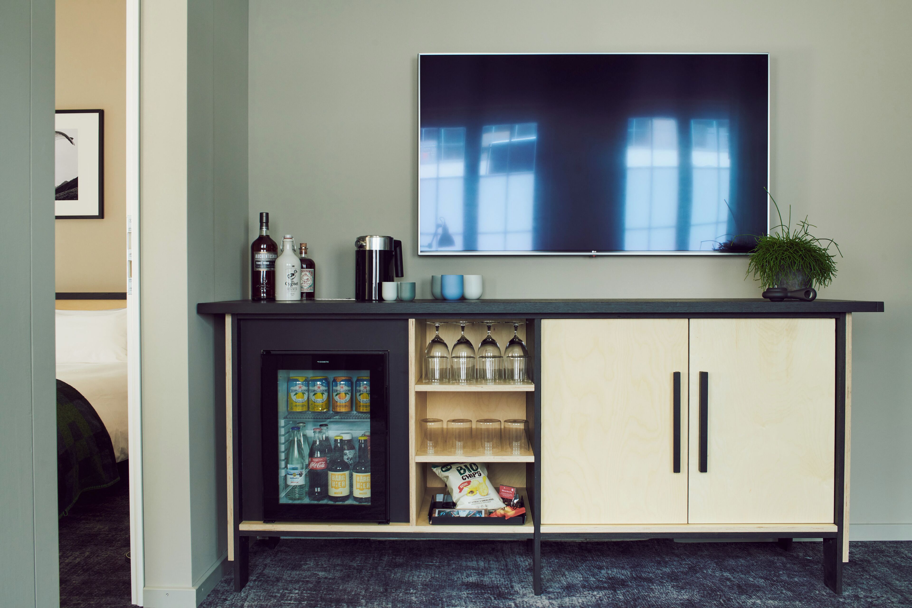 A minibar with a refrigerator, shelves with bottles, glasses, and snacks; a flat-screen TV above; and a potted plant on the right.