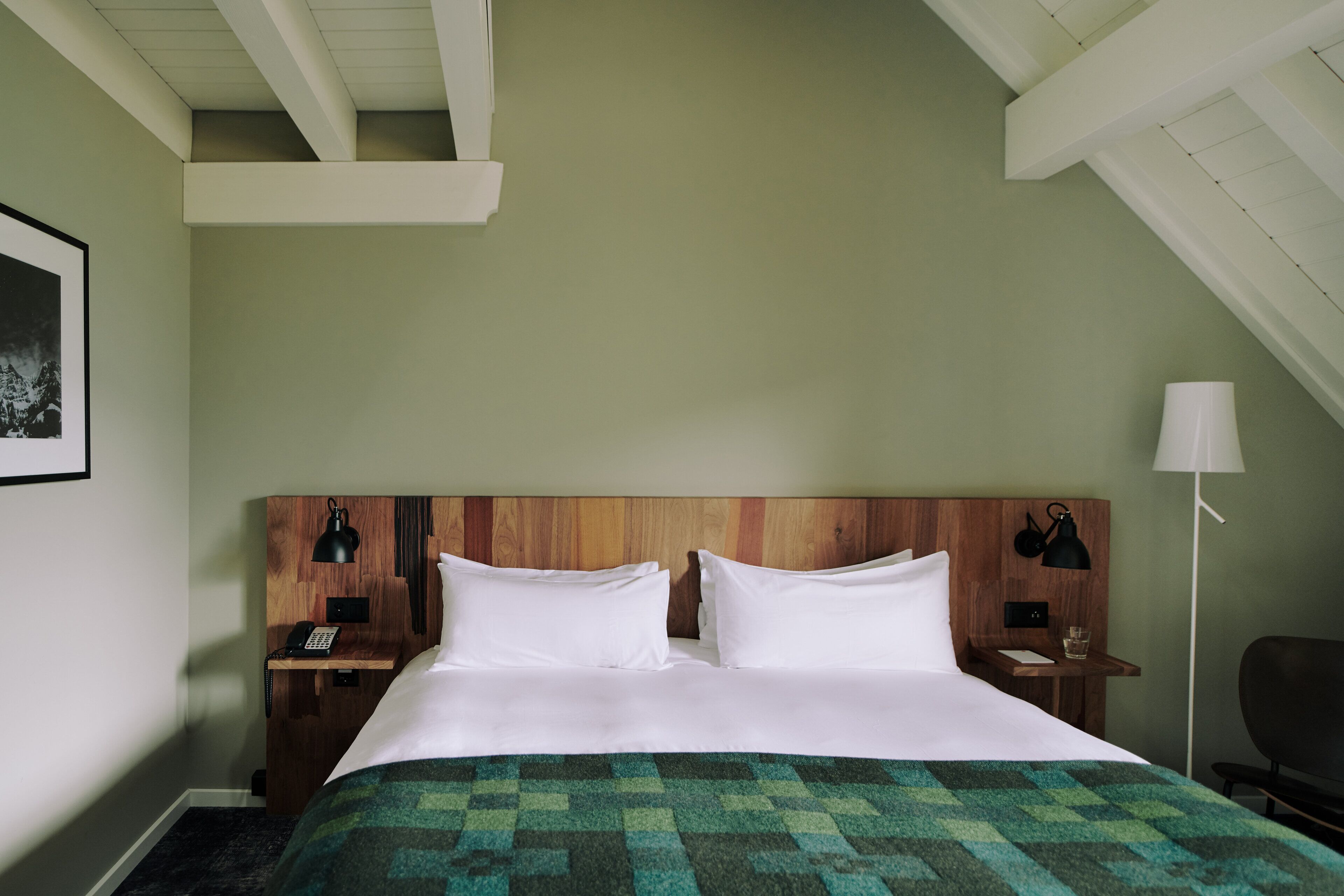 Una cama bien hecha con colcha verde estampada, cabecera de madera, lámparas de noche y mesita de noche con teléfono y vaso.