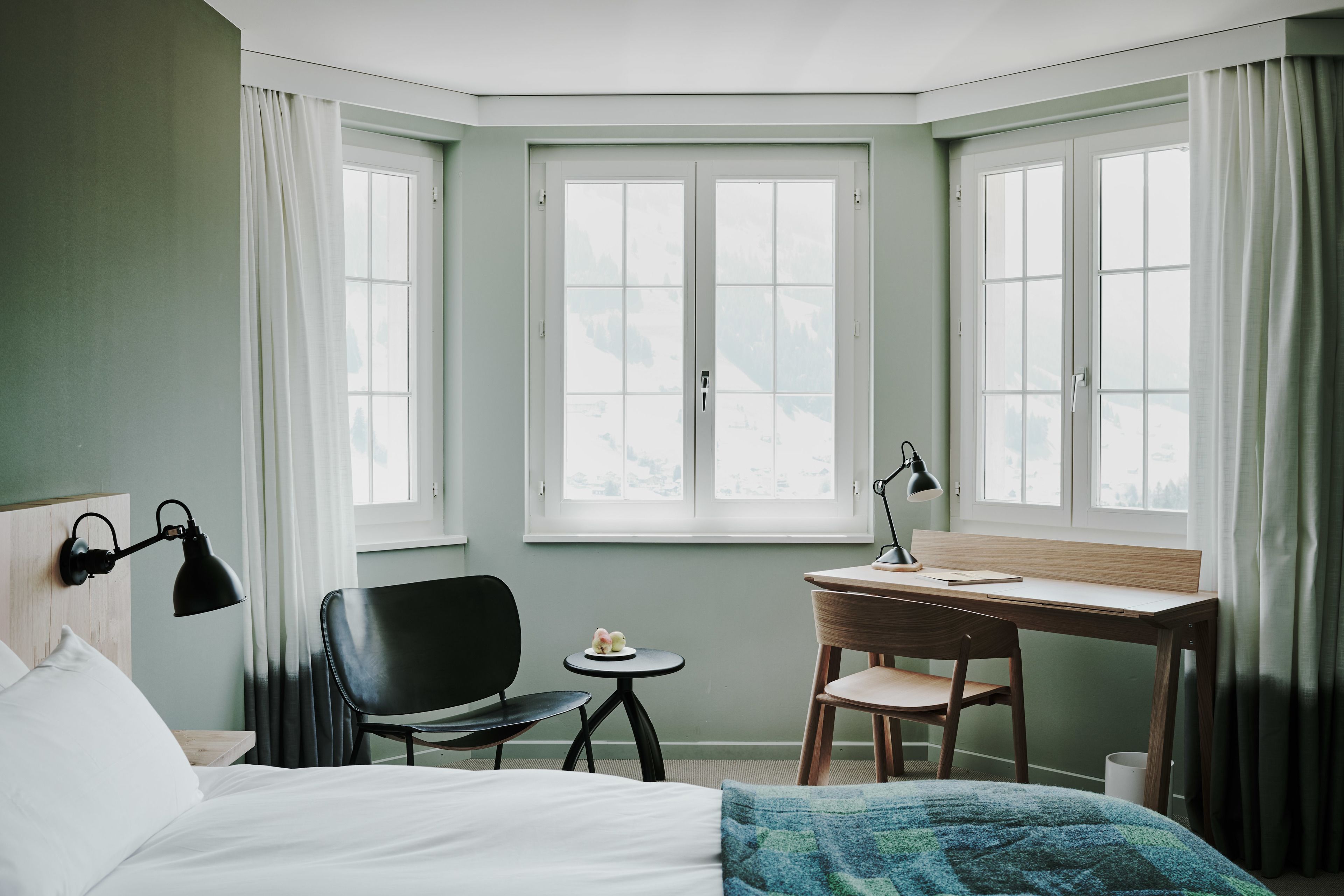 Hotellihuone, jossa on sänky, työpöytä, tuoli, pieni pöytä ja ikkuna, jossa on verhot.