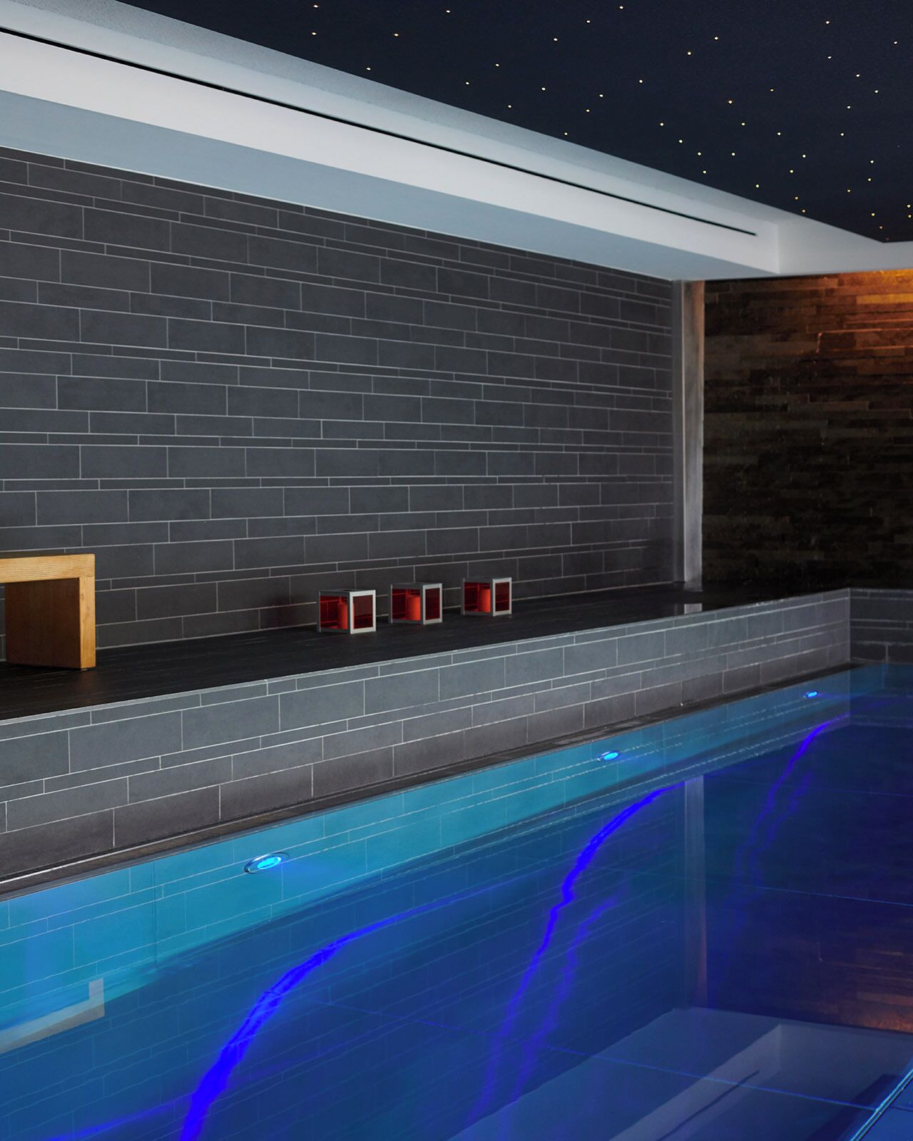A modern indoor pool area with a dark tiled wall, a wooden bench, and a red-lit shelf.
