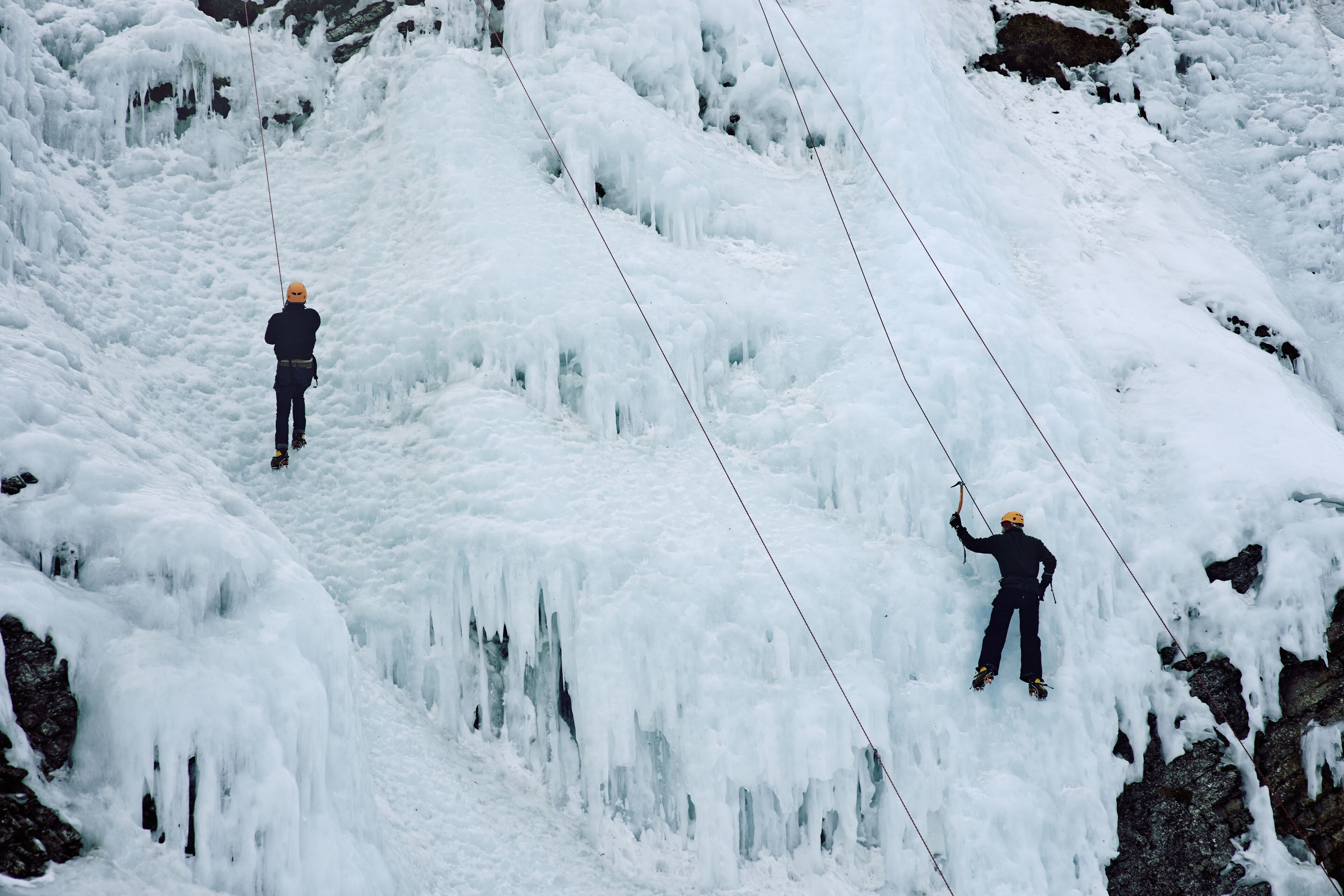 Two individuals rappelling down a snowy slope with ice formations.
