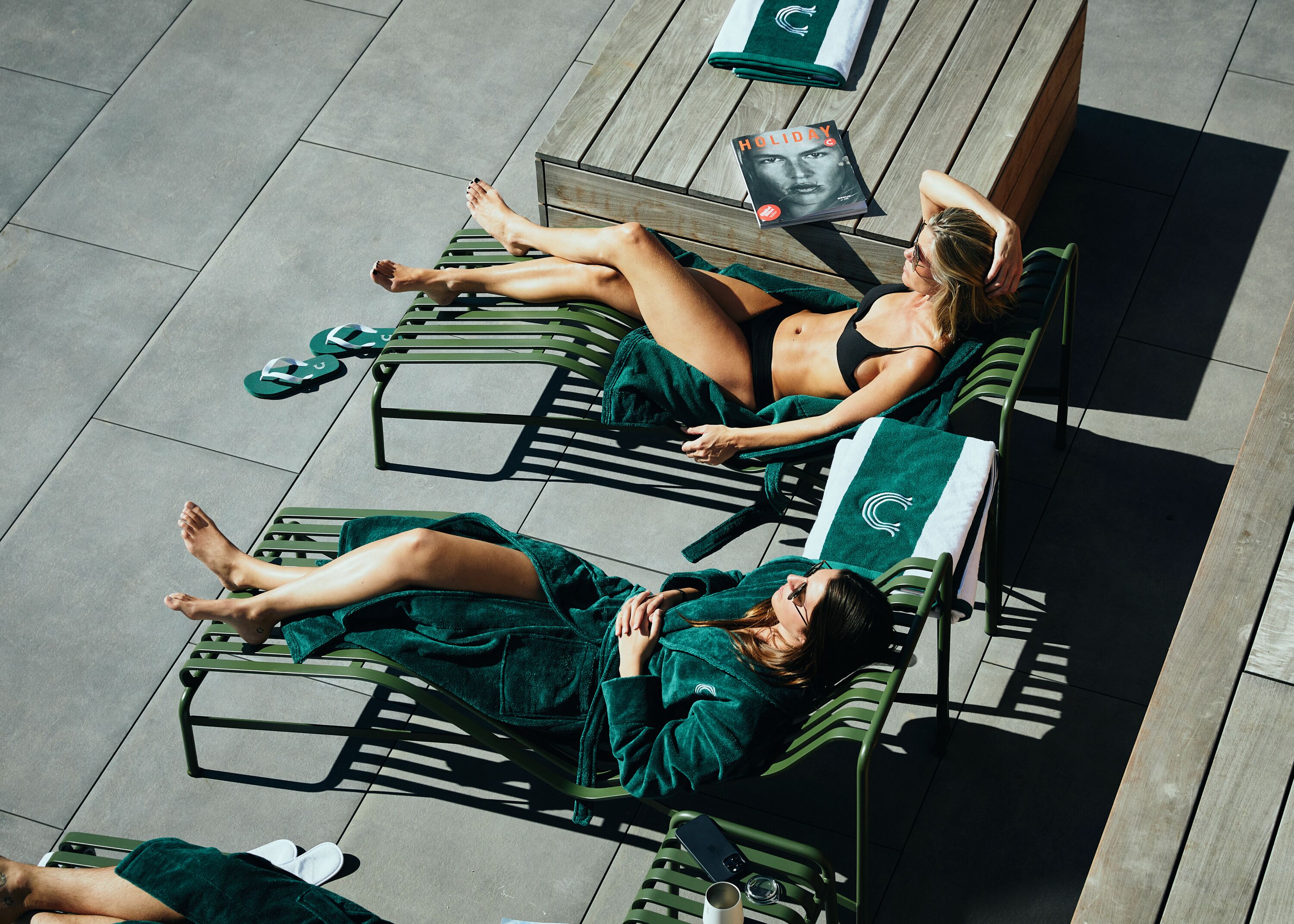 Two people sunbathing on green lounge chairs with a book titled "Holiday" beside them.
