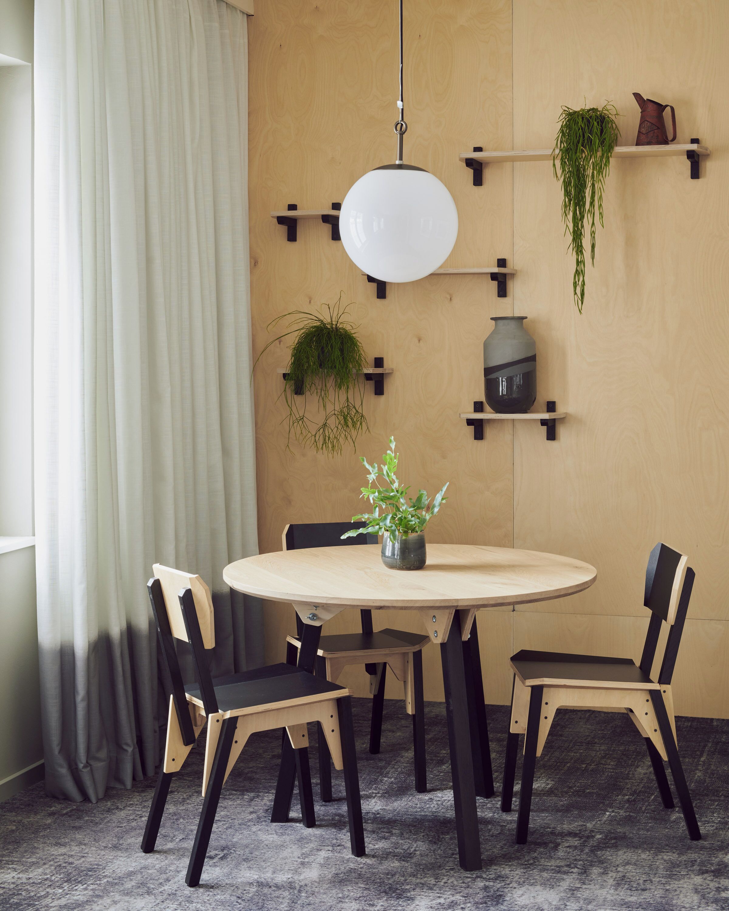 A dining area with a round table, six chairs, a hanging light fixture, and wall-mounted shelves with plants and a vase.