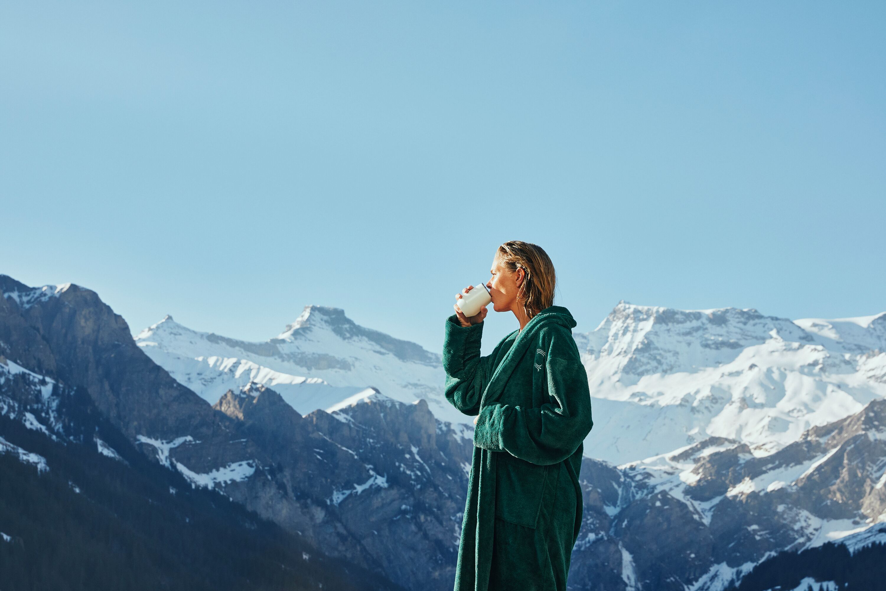 Una persona vestida con una bata verde, de pie en una zona montañosa con picos nevados al fondo.