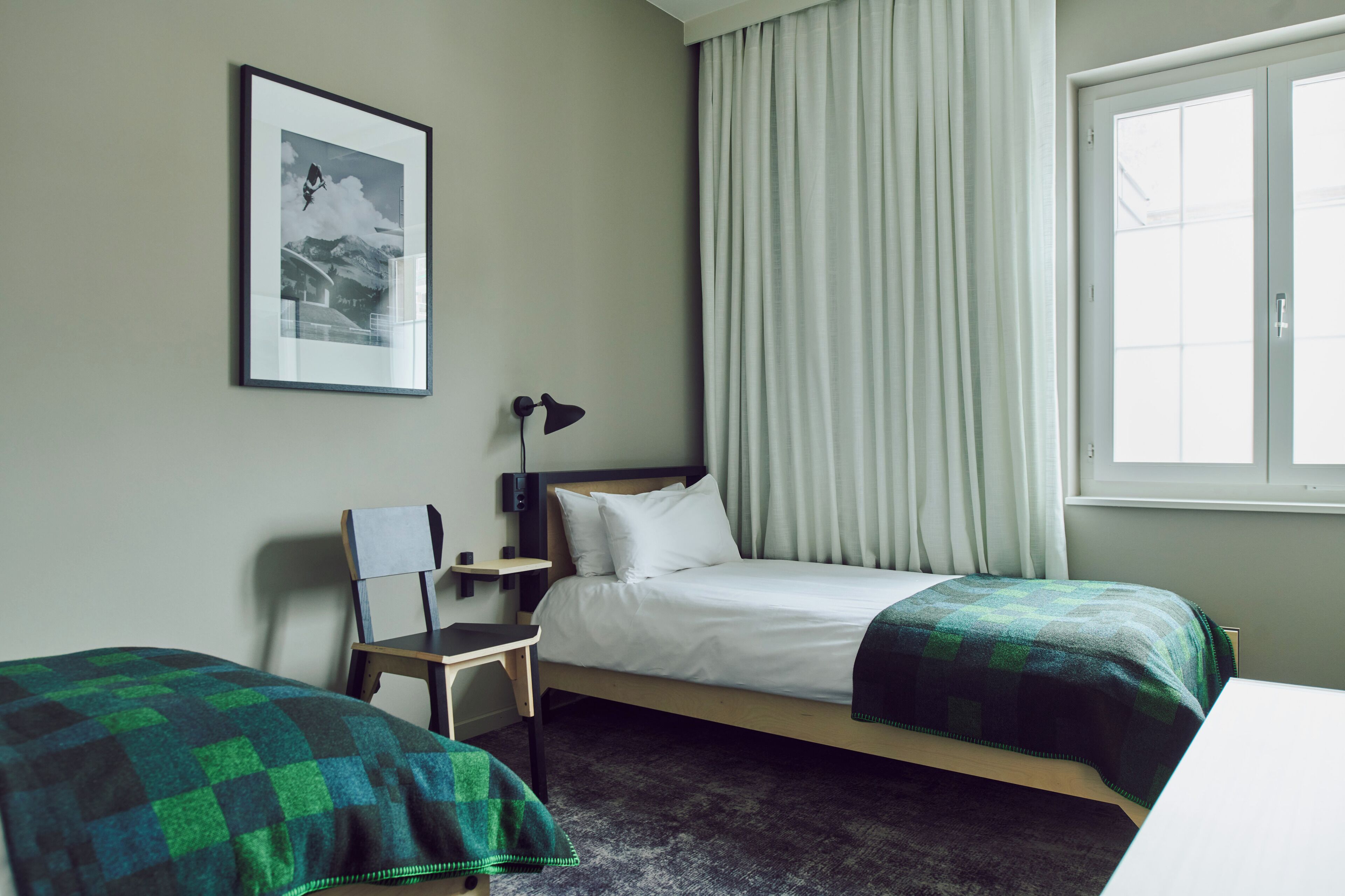 Une chambre d’hôtel avec un lit, deux tables de chevet, une chaise, un bureau, une lampe, un tableau encadré au mur et une fenêtre avec des rideaux.