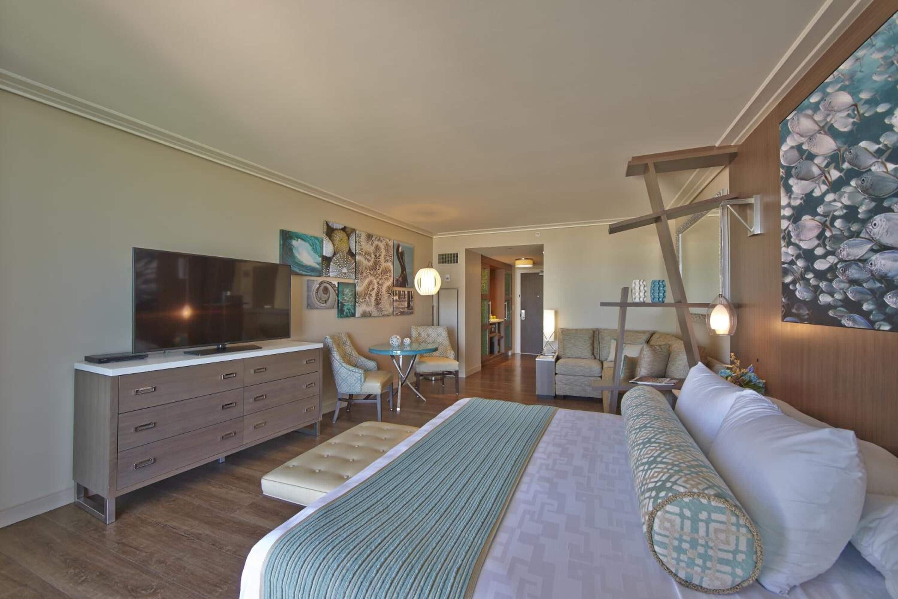 A modern hotel room with a large bed, a TV, a desk, and a dining area.