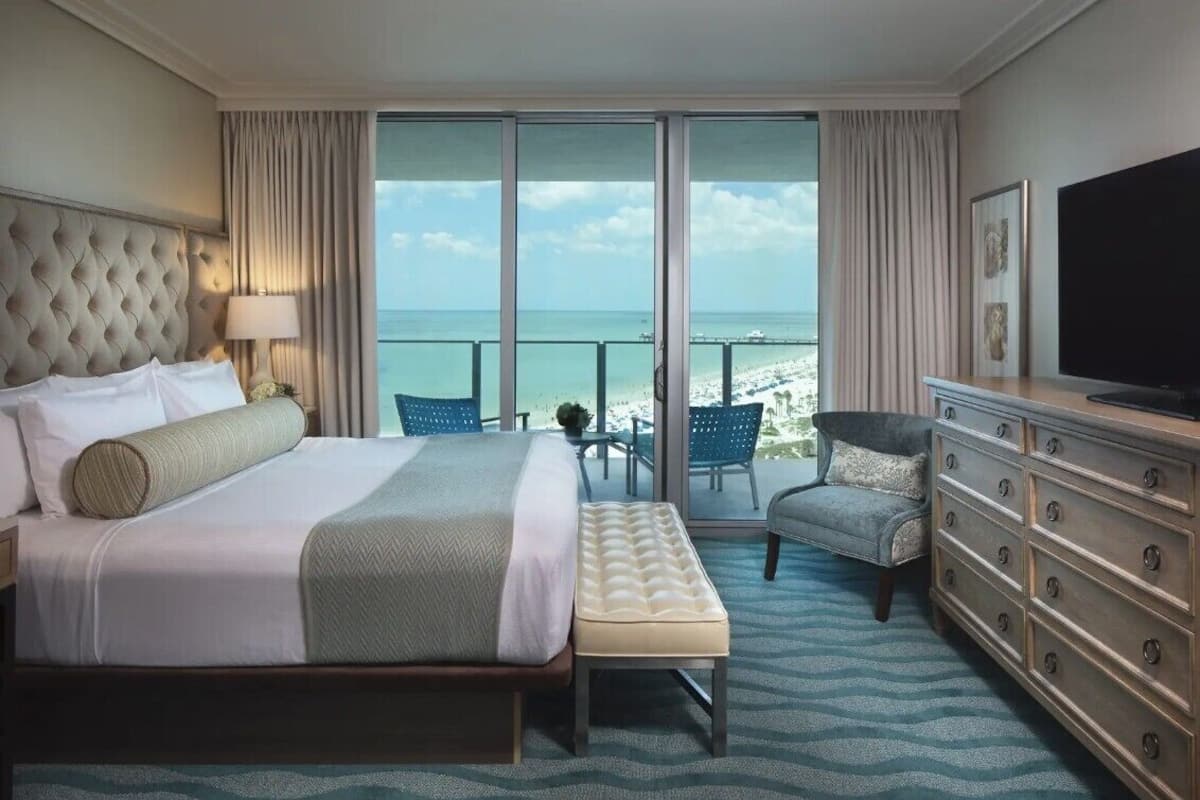 A hotel room with a large bed, a TV, a dresser, and a balcony with a view of the beach.