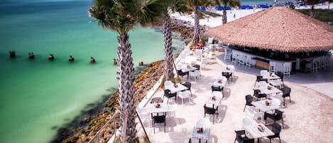 An outdoor seating area with tables and chairs, a thatched-roof structure, and a view of the beach and ocean.