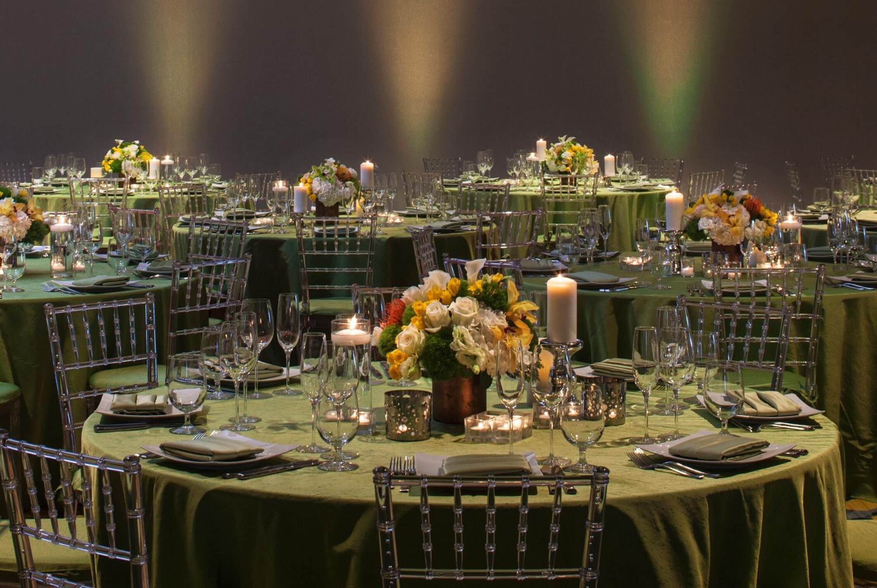 A formal dining setup with multiple tables, each set with green tablecloths, candles, and floral centerpieces.