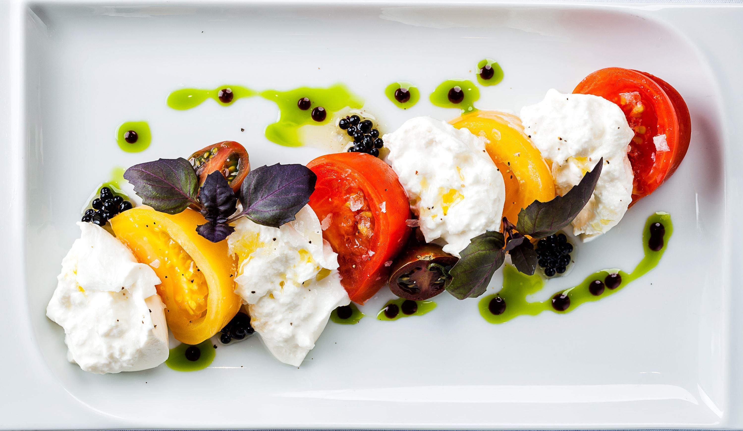 A white plate with a dish of tomatoes, mozzarella, and blackberries, garnished with green sauce.