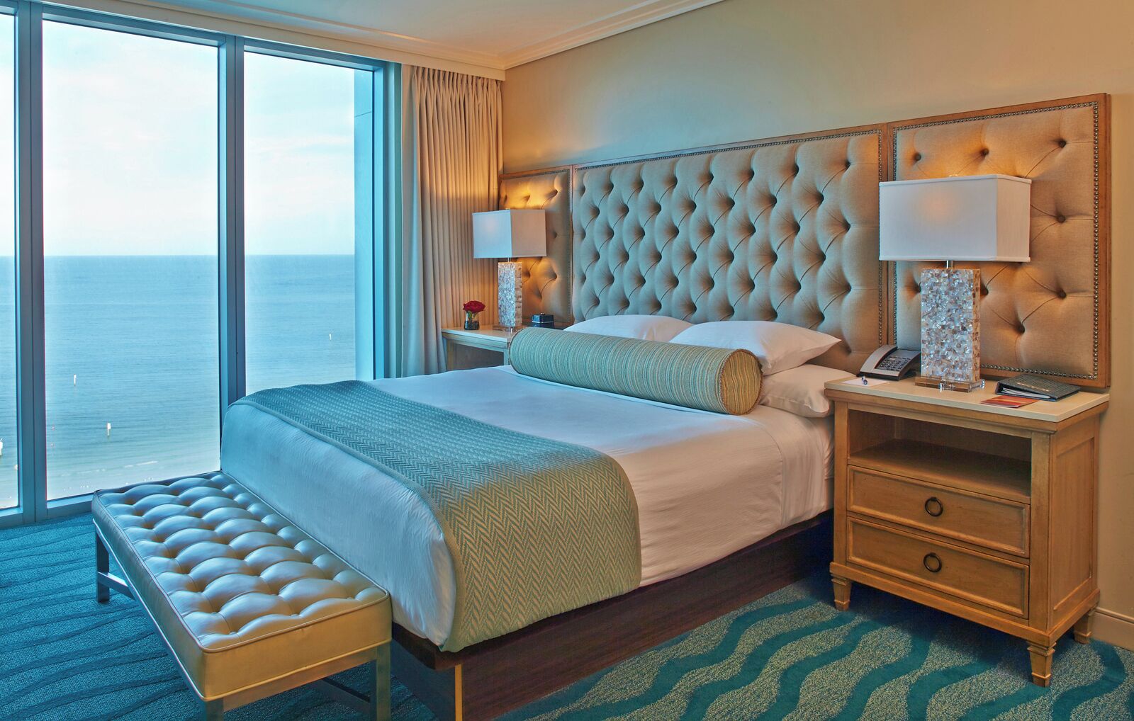 A hotel room with a large bed, a bench, a nightstand, and a view of the ocean.