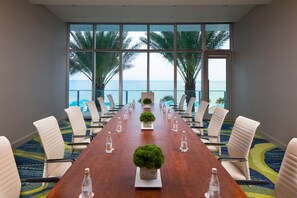 A conference room with a long table, white chairs, and water bottles. It has large windows with a view of palm trees and the sea.
