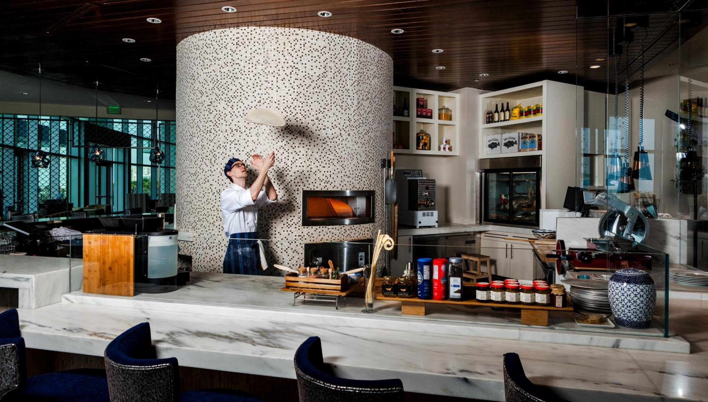 A modern bar area with a chef preparing drinks, a large textured column, and a variety of bottles and jars on shelves.