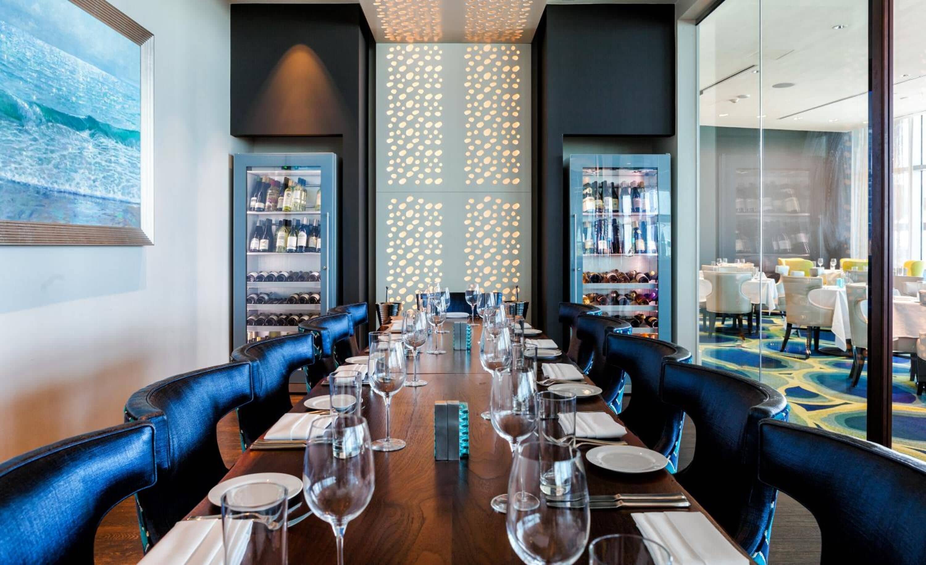 A dining area with a long table set for a meal, flanked by wine racks and a cityscape view.