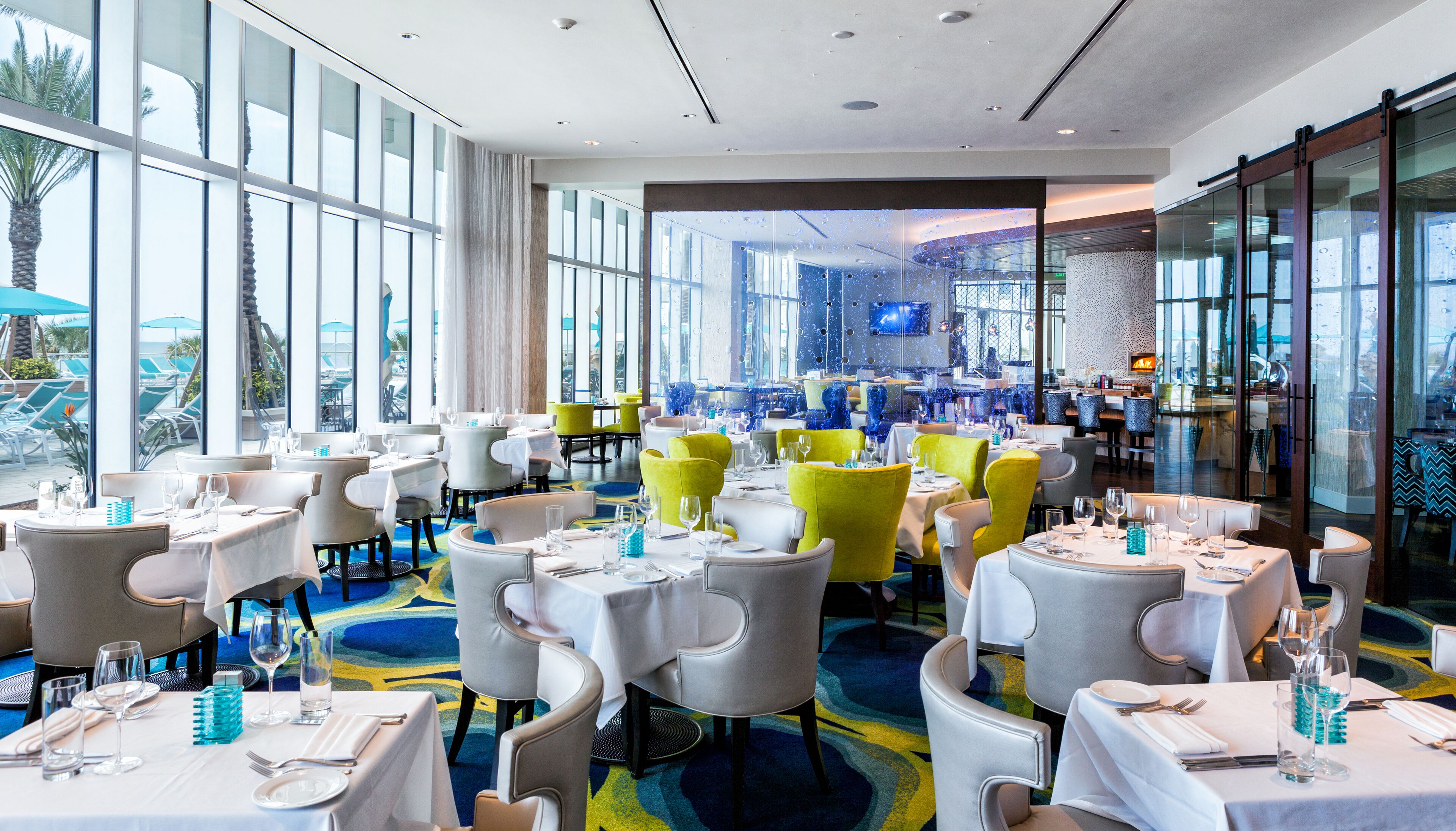 A modern dining area with tables set for guests, large windows offering views of palm trees, and a vibrant carpet design.