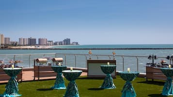 A rooftop terrace with tables set up for dining, overlooking a beach and city skyline.
