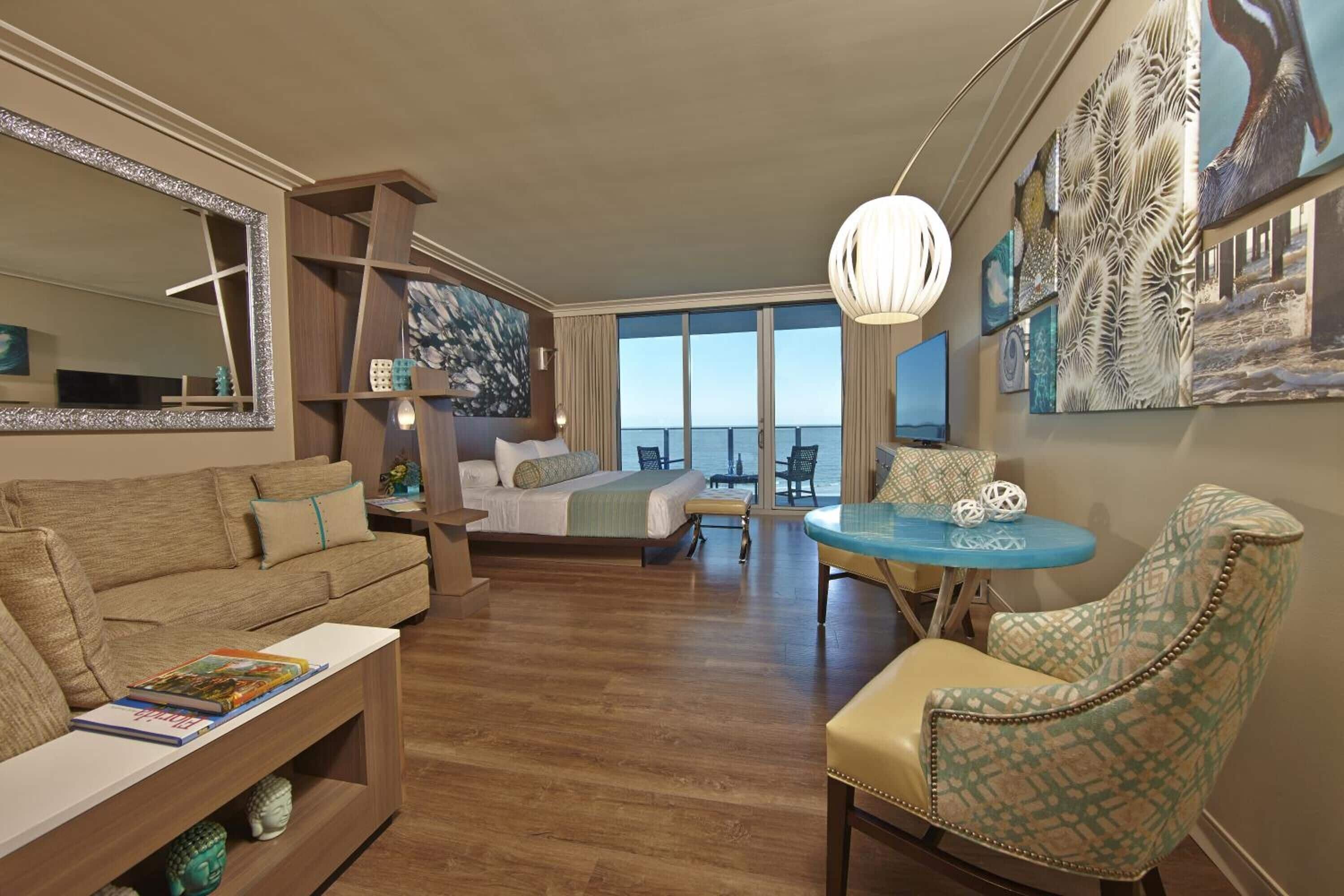 A modern hotel room with a large bed, a sofa, a dining table, and a view of the sea.