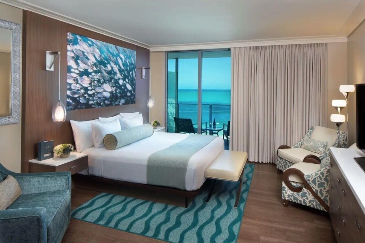 A hotel room with a large bed, a desk, a chair, a TV, and a balcony with a view of the ocean.