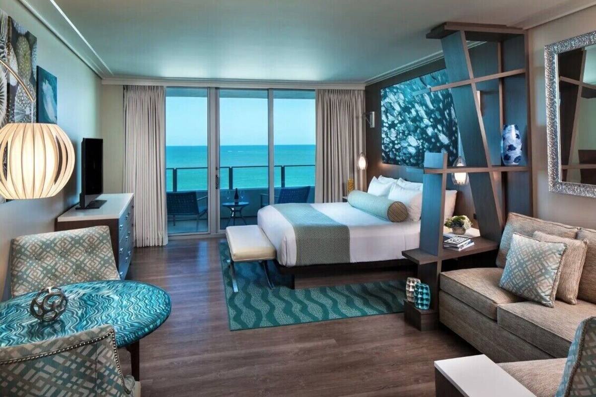 A modern hotel room with a large bed, a sofa, a desk, and a view of the ocean.