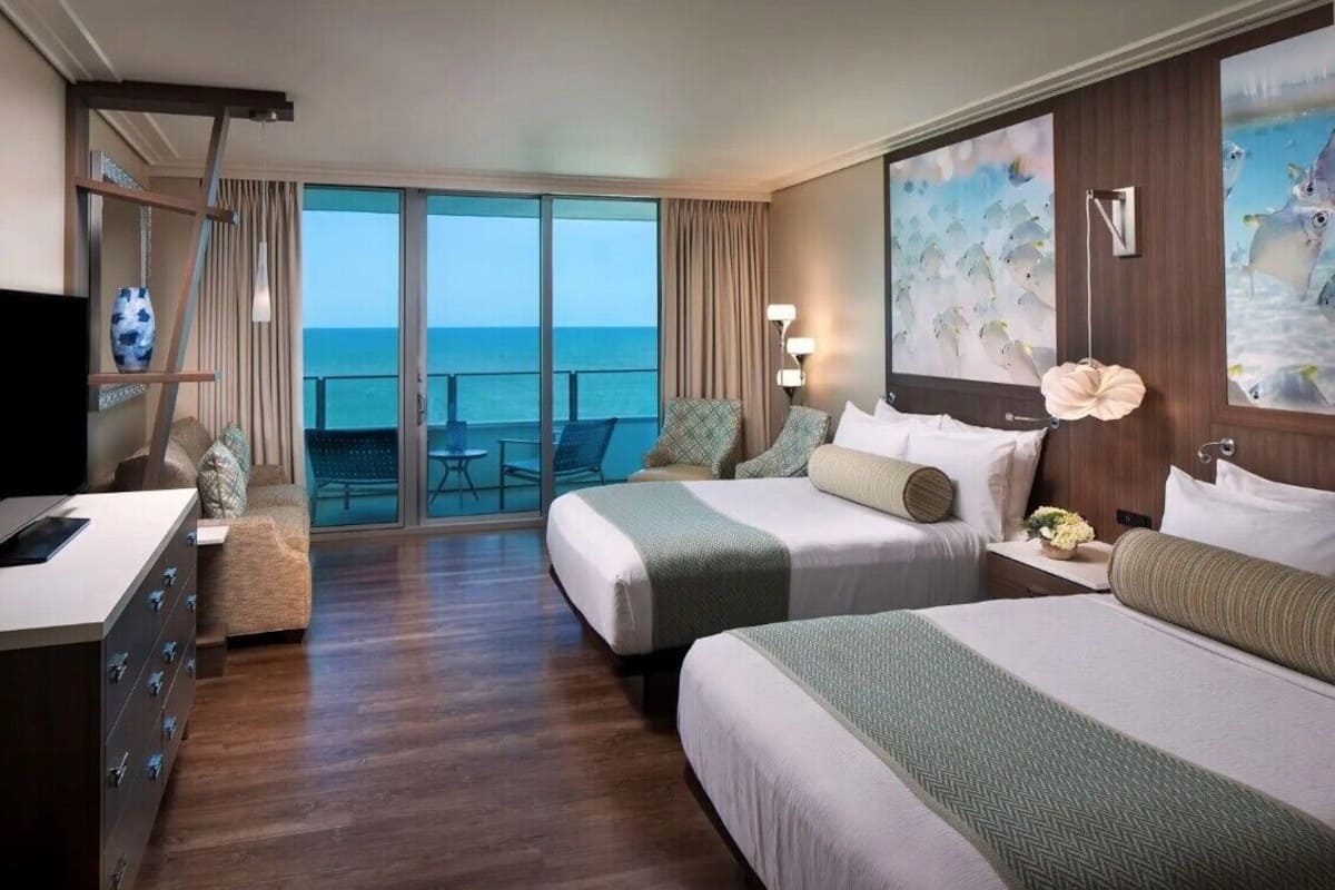 A hotel room with two beds, a desk, and a balcony with a view of the sea.
