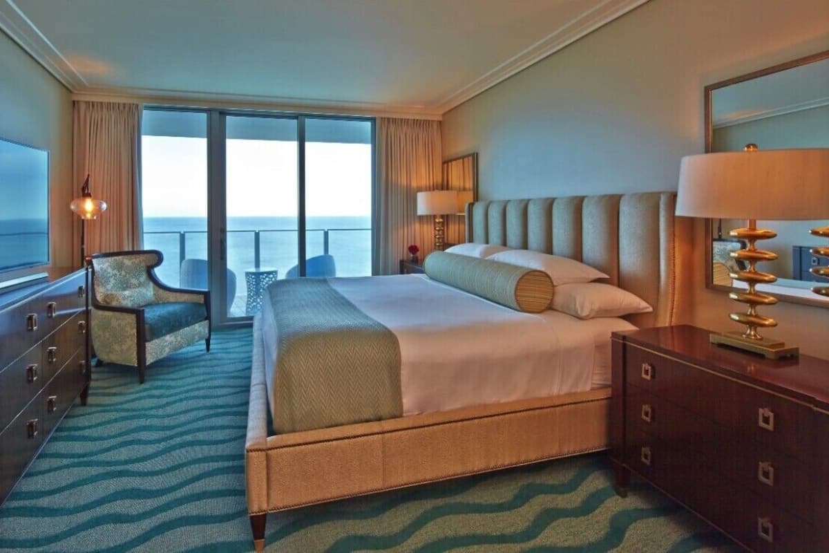 A hotel room with a large bed, a television, a chair, a dresser, and a view of the ocean.