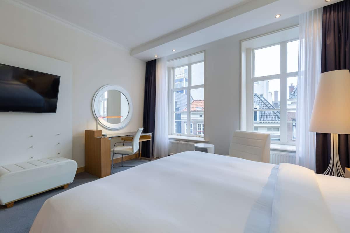 A hotel room with a large bed, a desk with a chair, a round mirror, and a TV mounted on the wall.