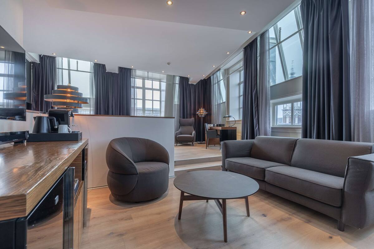 A modern living room with a sofa, armchair, and coffee table. There is a kitchen area with a counter and appliances. Large windows with curtains are visible.