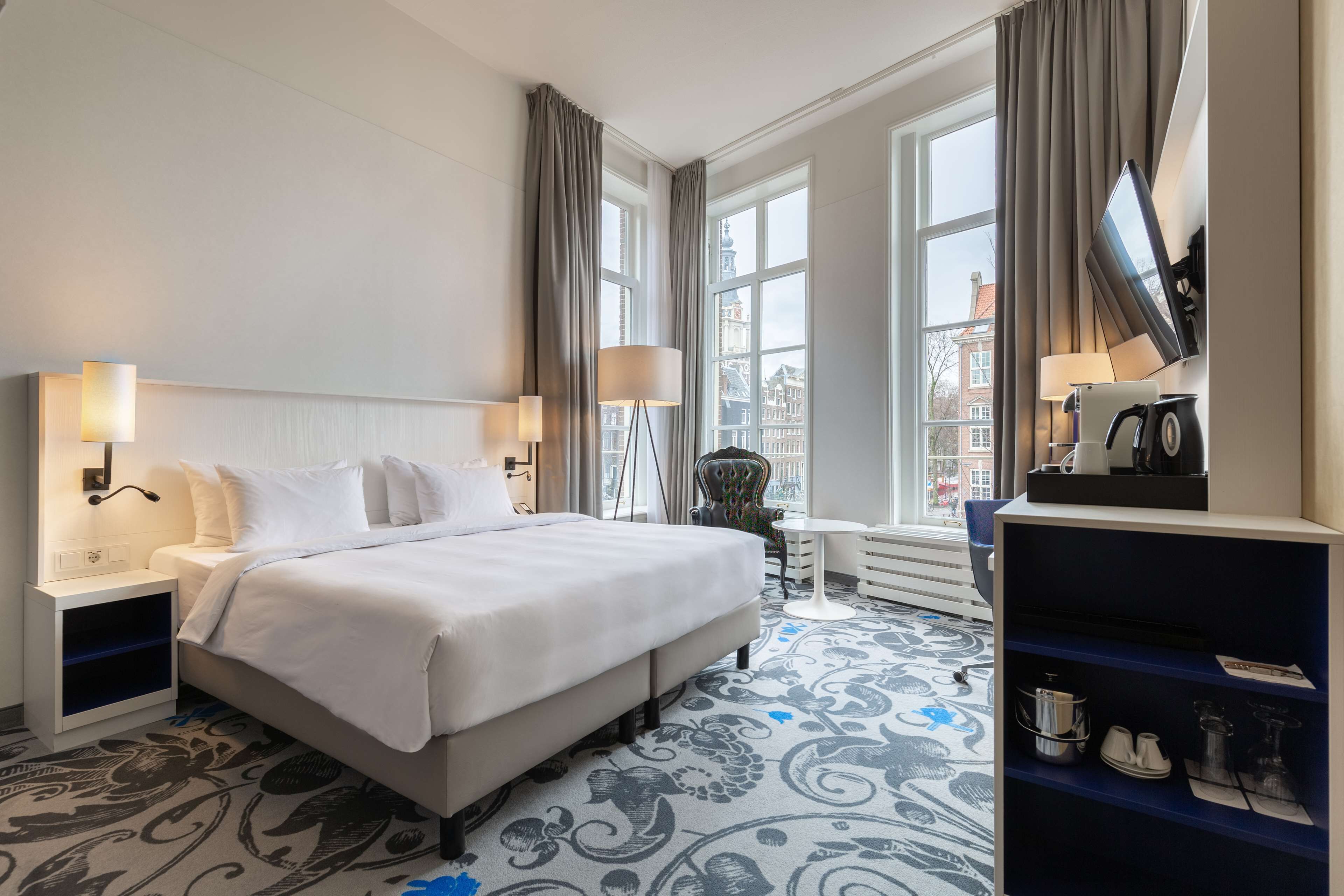 A hotel room with a large bed, two bedside tables with lamps, a TV, a small table with a chair, and a bookshelf.