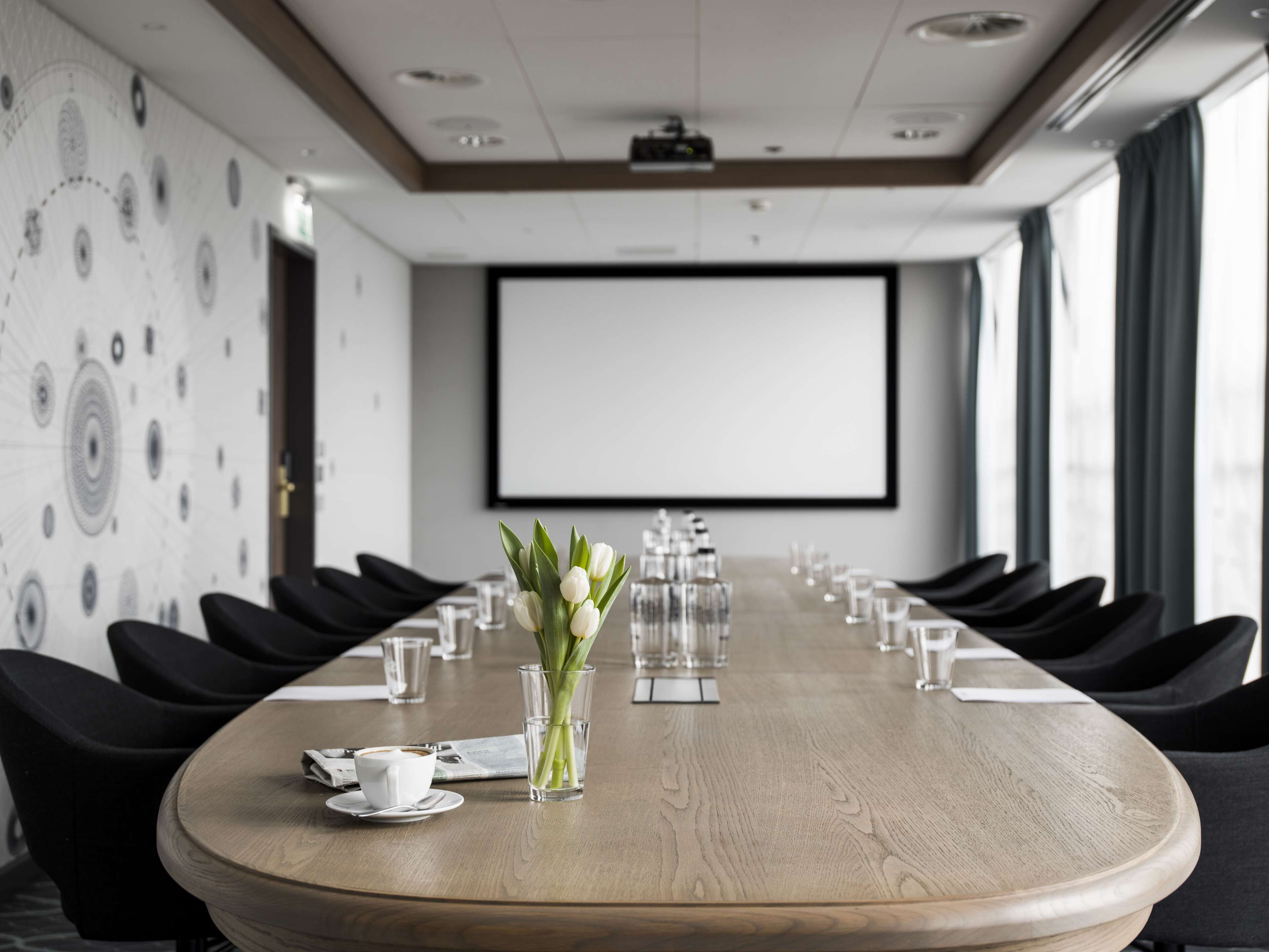 Un salón de conferencias con una larga mesa, sillas, una pantalla de proyector y un arreglo floral.