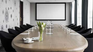 Un salón de conferencias con una larga mesa, sillas, una pantalla de proyector y un arreglo floral.