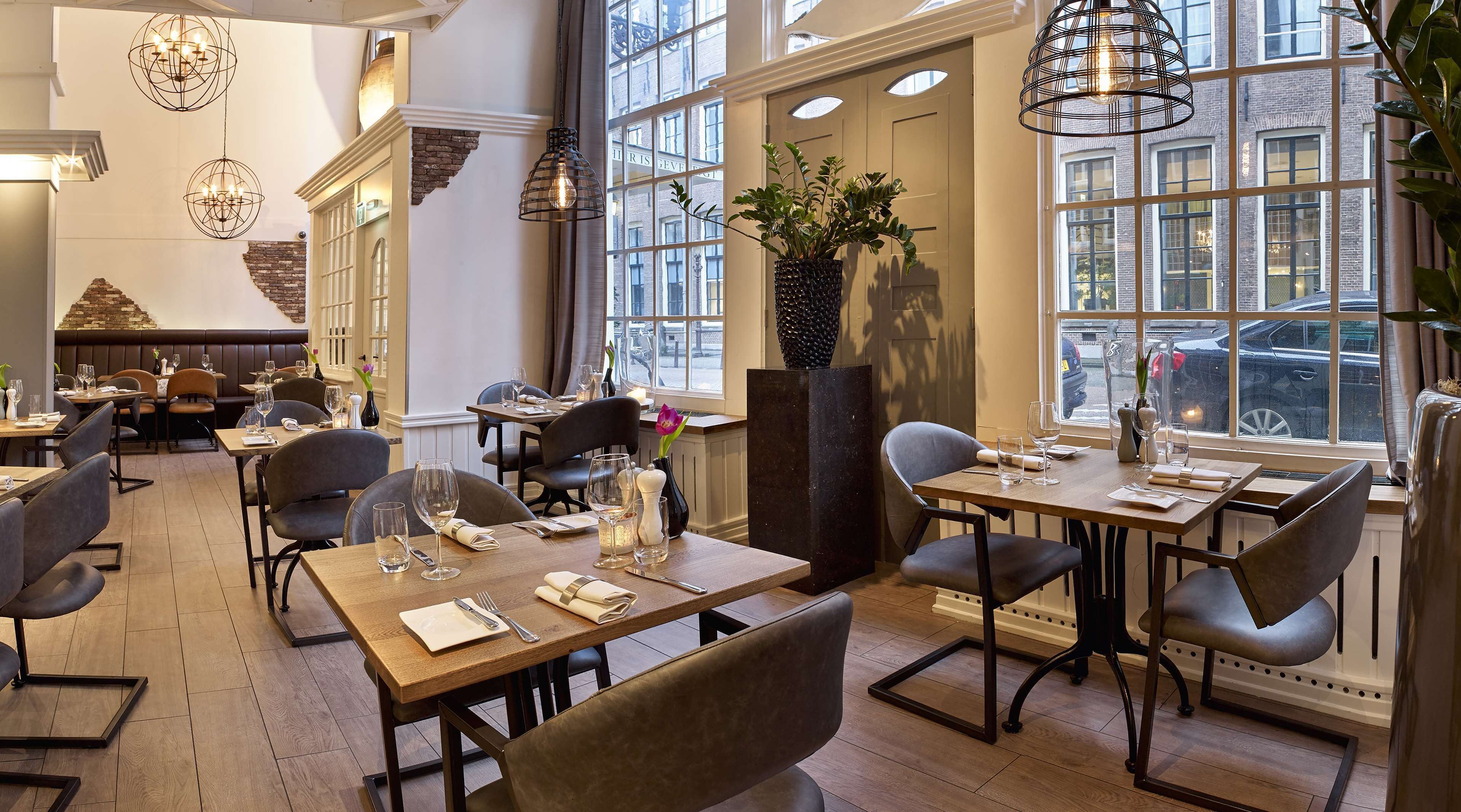 A restaurant with high ceilings, large windows, and modern lighting fixtures. Tables are set with wine glasses and cutlery.