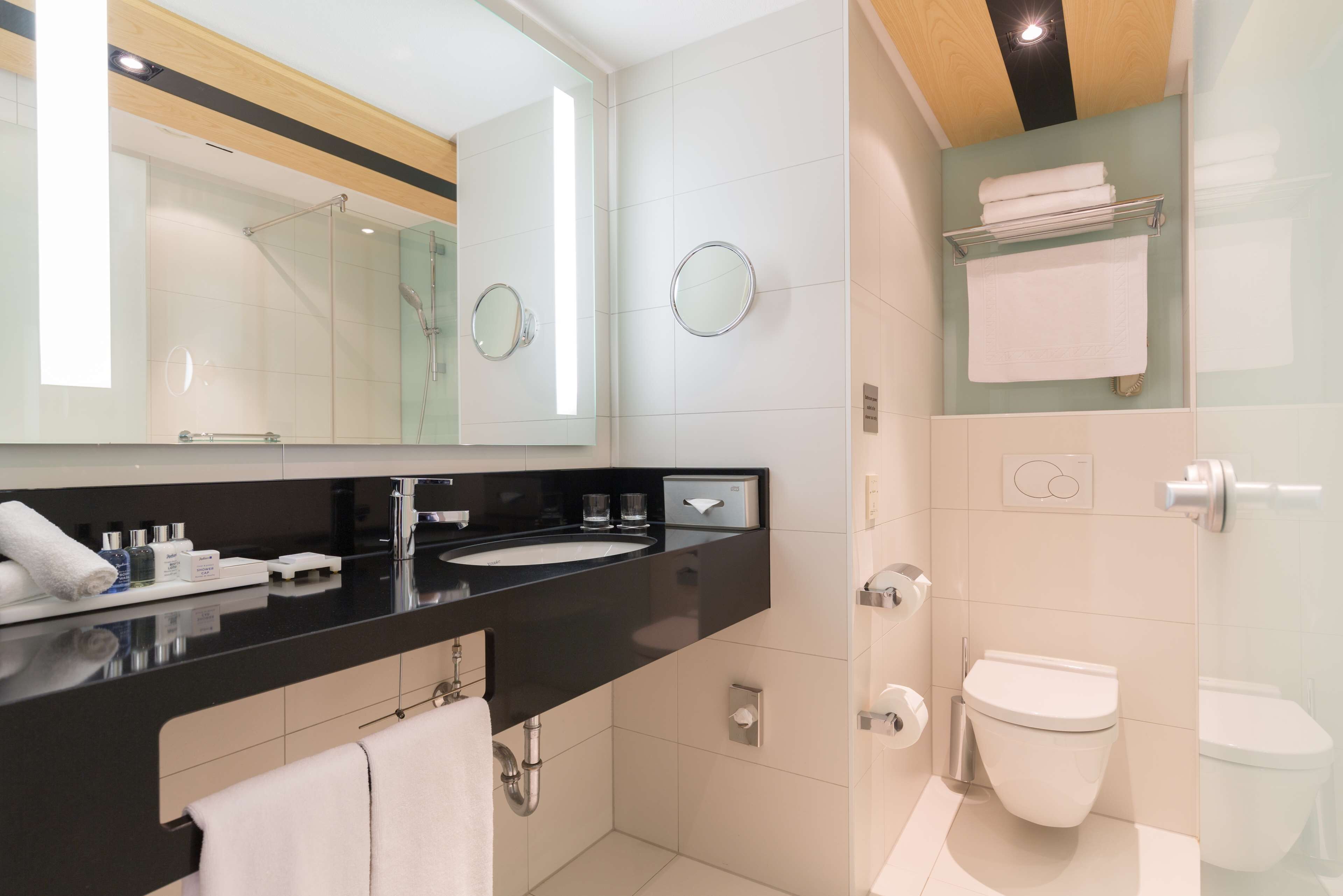 Un baño moderno con encimera negra, dos lavamanos, un espejo, cabina de ducha de vidrio, inodoro y toallero.