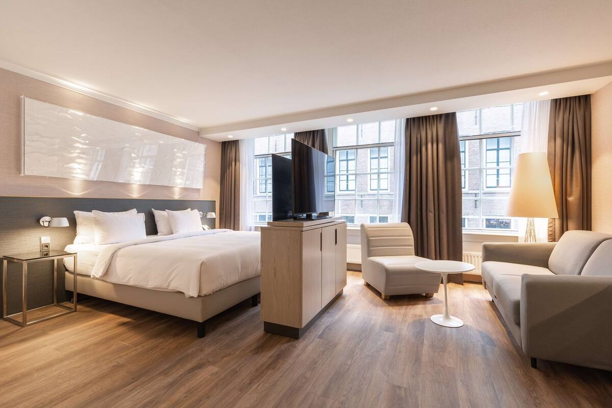 A modern hotel room with a bed, a desk, a TV, a sofa, and a lamp.