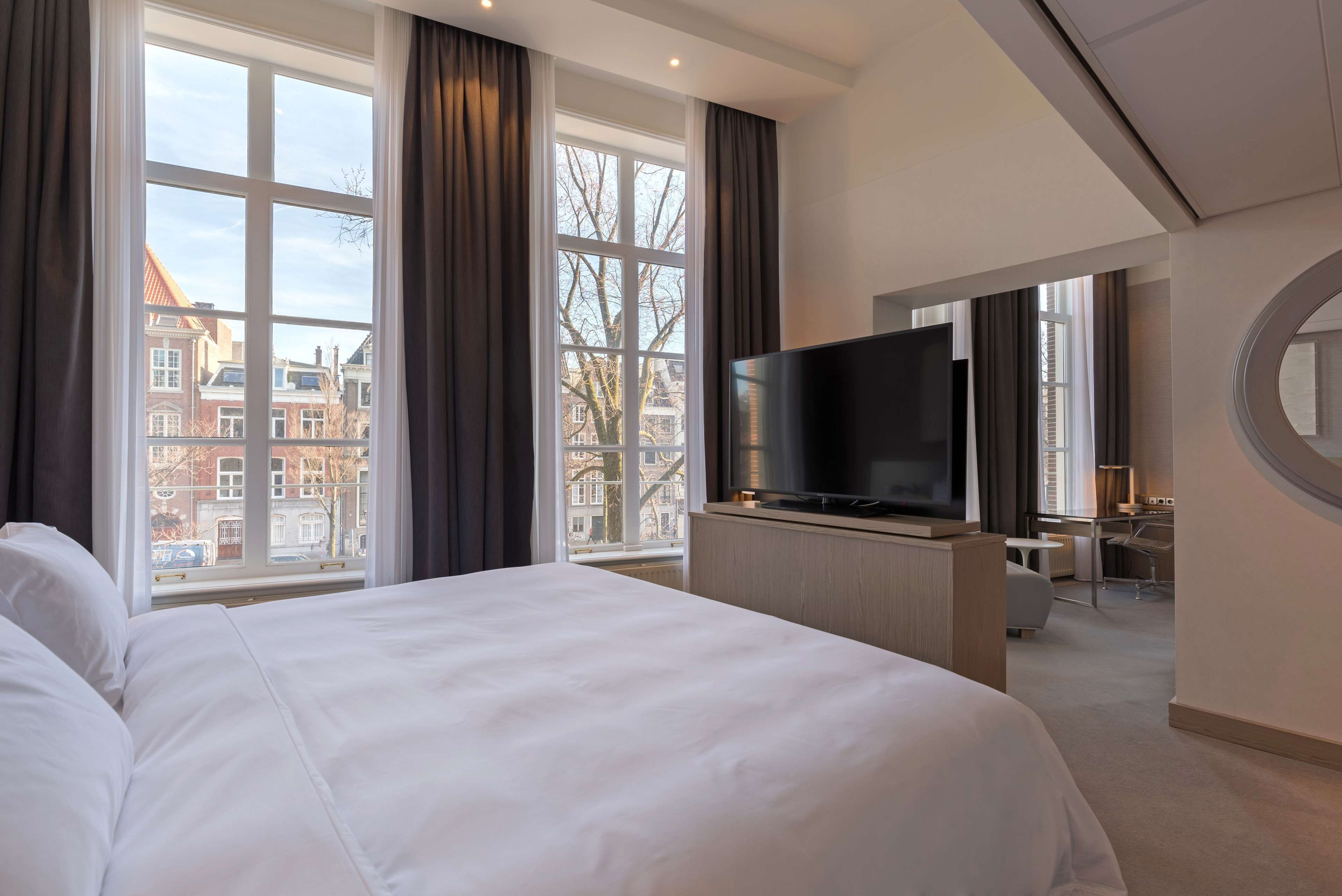 A hotel room with a large bed, a flat-screen TV, a desk, and a view of buildings through large windows.