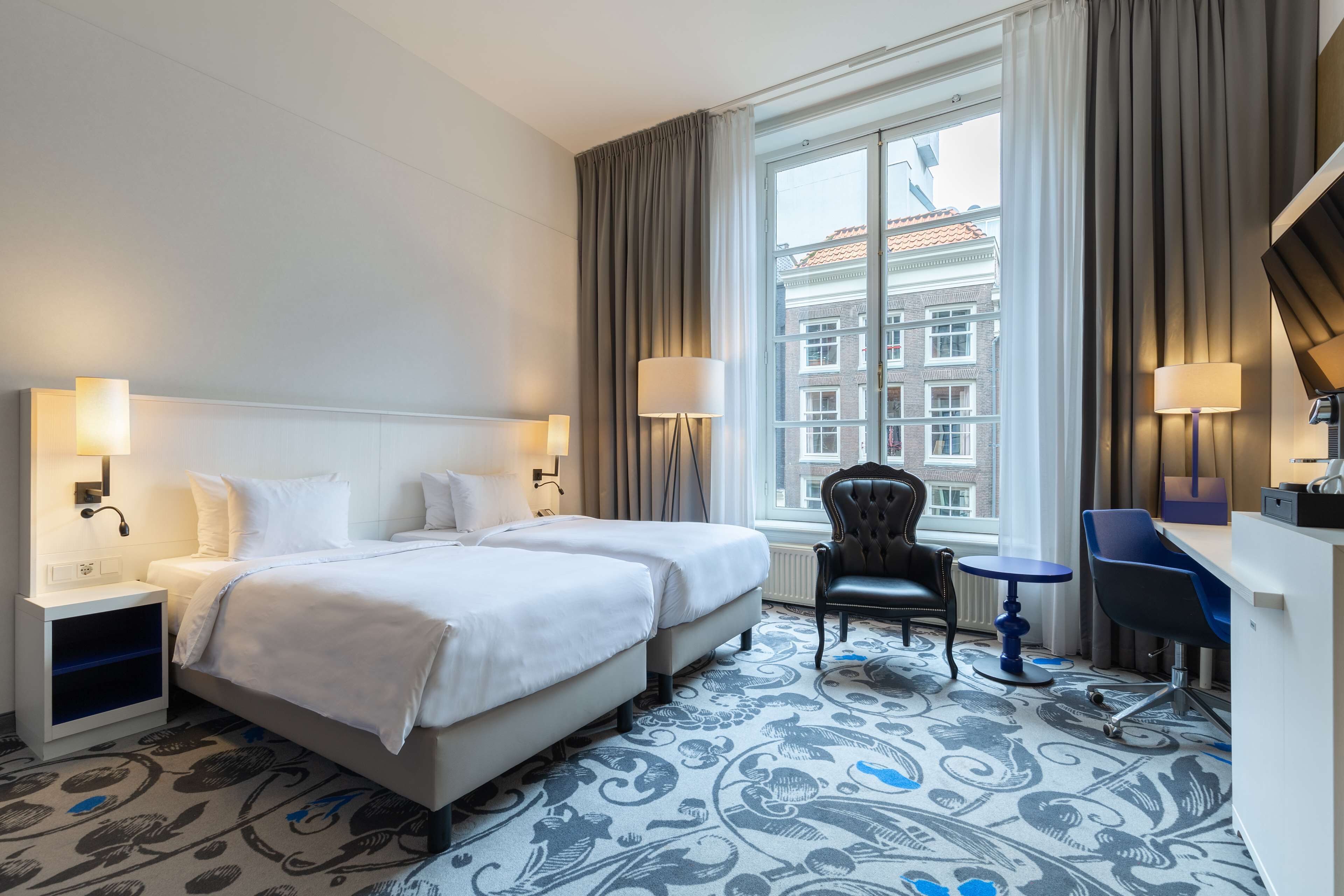 A hotel room with two beds, a desk, a chair, and a view of buildings through the window.