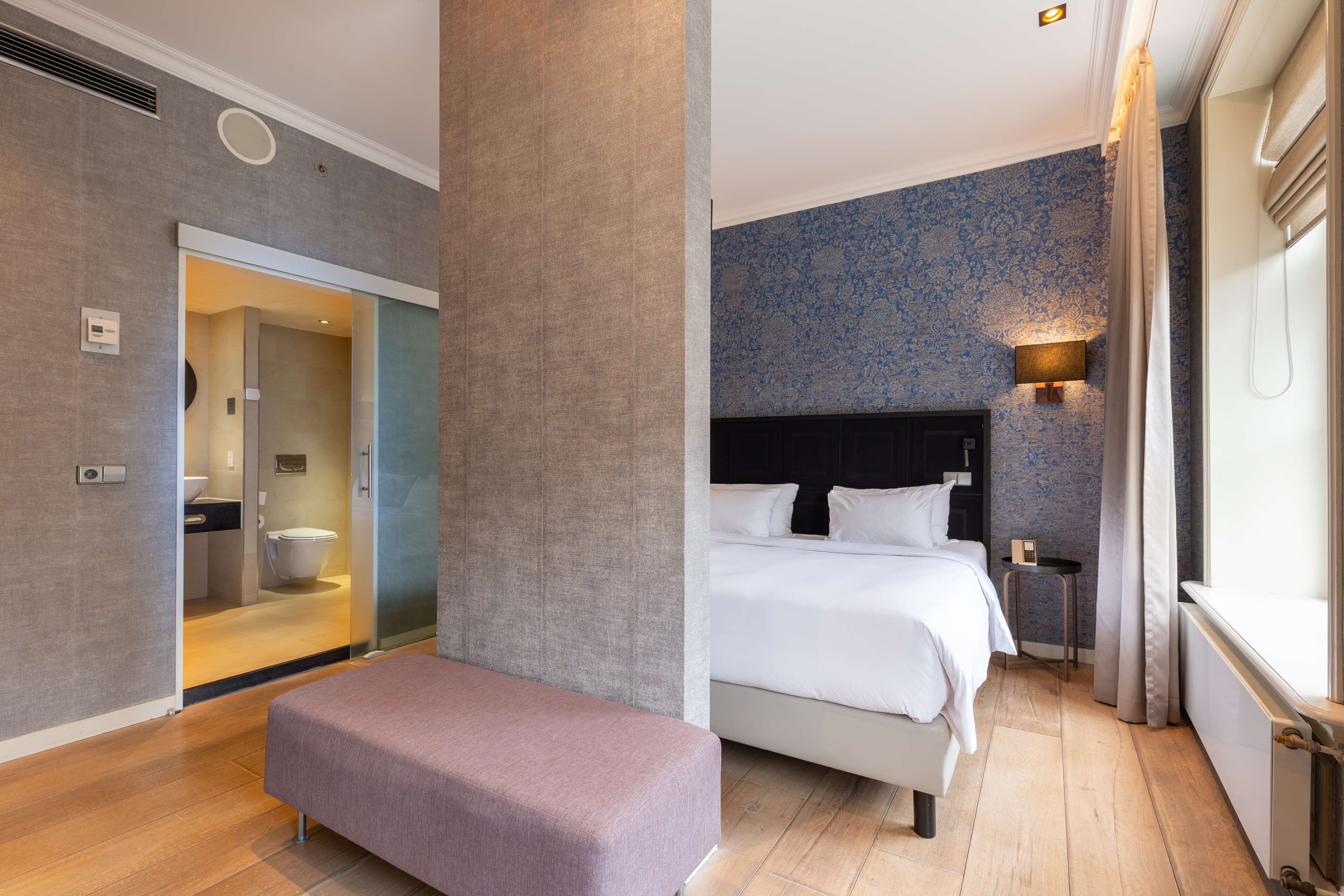 Une chambre d’hôtel moderne avec un grand lit, un banc, une petite table et une salle de bain visible à travers une porte en verre.