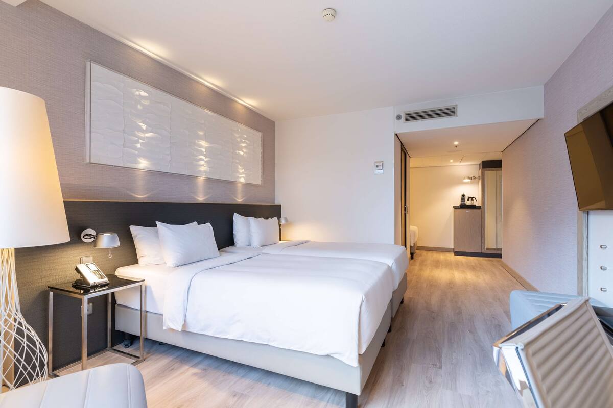 A modern hotel room with a large bed, bedside table with a lamp, a phone, and a wall-mounted artwork.