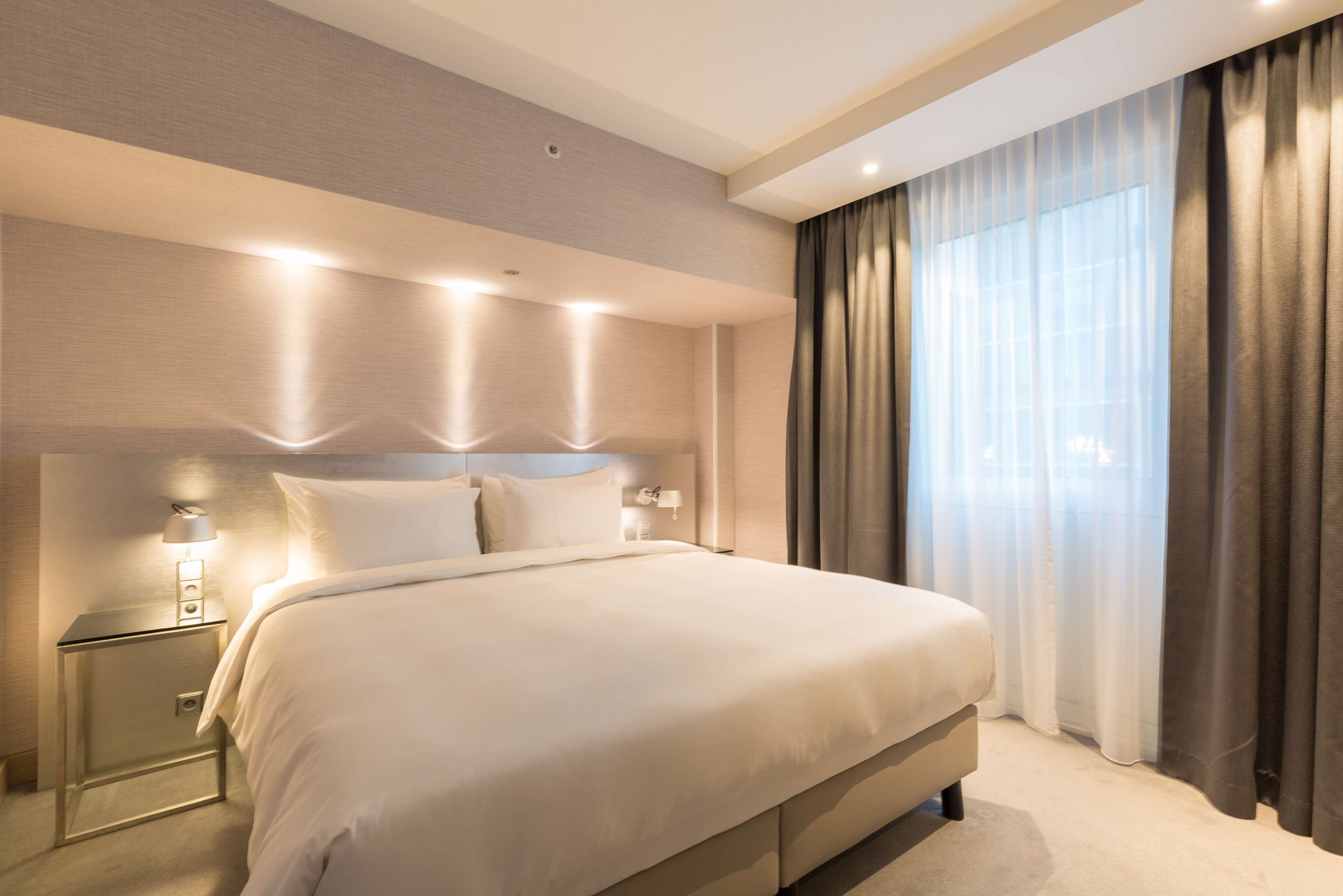 A modern hotel room with a large bed, a nightstand with a lamp, and a view of the city through sheer curtains.