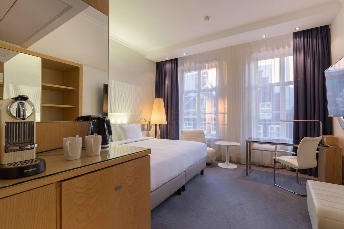 A hotel room with a bed, a desk, a chair, a coffee maker, and a lamp.