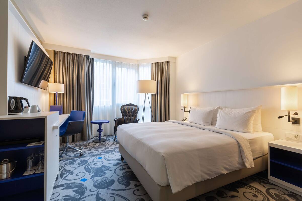 A hotel room with a bed, two bedside tables, a desk with a chair, a TV mounted on the wall, and a window with curtains.